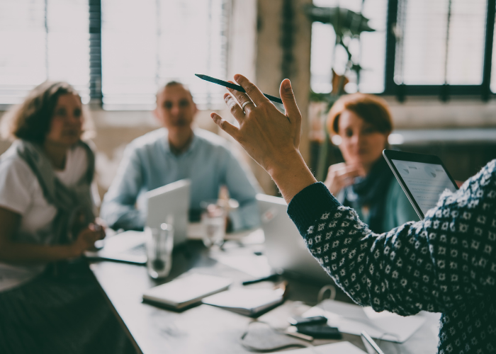 three Employees at a table with laptops getting instruction from a supervisor, of whom we only see her arm and hand